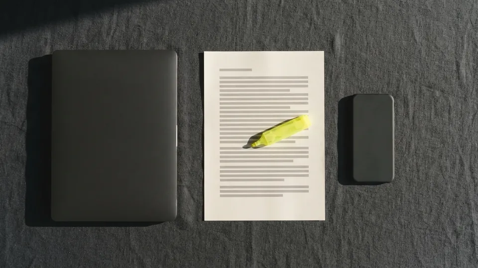 A closed laptop, a printed page with a yellow highlighter lying on it and a face-down phone laid out in a row