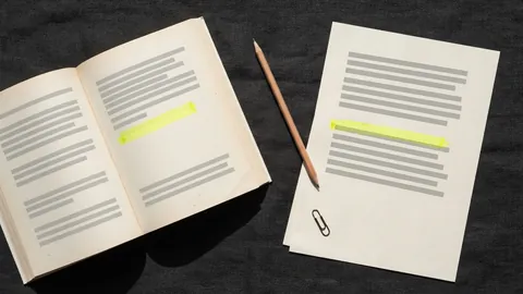 An open book and an essay sheet on a charcoal desk, the same short passage highlighted in yellow on both, a pencil between them