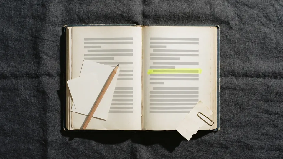 An old clothbound book open on a desk with one line highlighted in yellow and a blank index card and pencil resting on the page