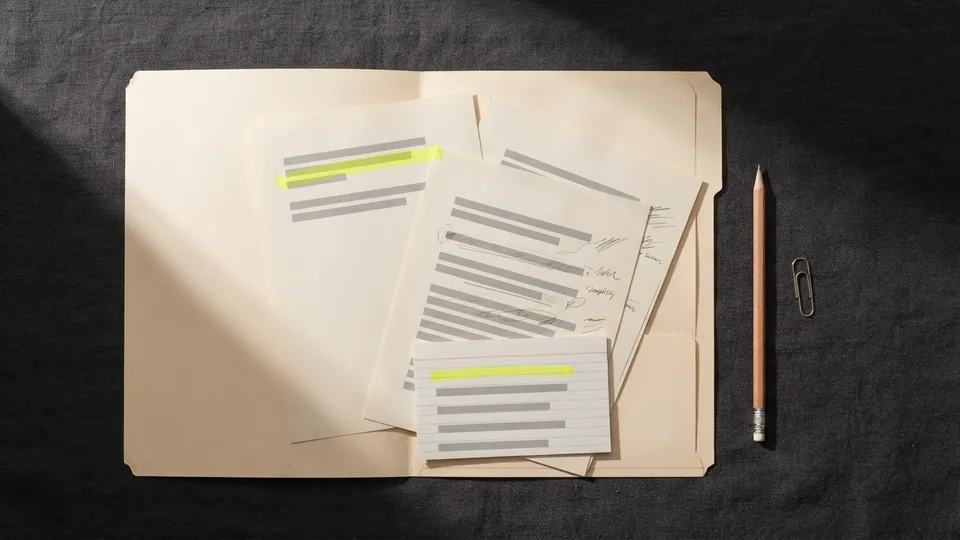 An open folder of drafts, a highlighted source sheet and an index card, the evidence record for one essay, on a charcoal desk