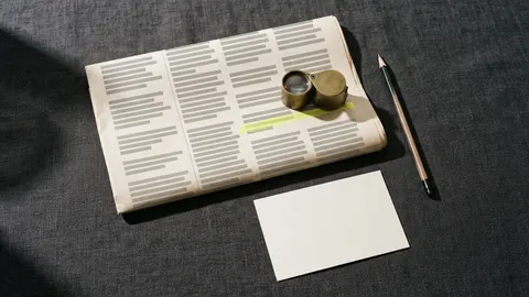A folded old newspaper with one line in a column highlighted in yellow, a loupe resting on it and a blank poster card below