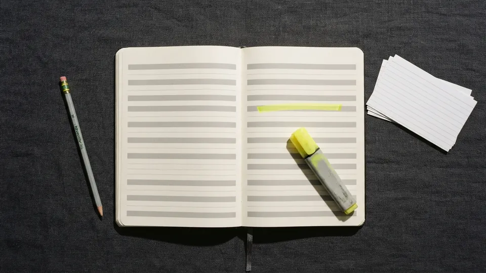 A ruled reading log notebook open on a charcoal desk, one row highlighted in yellow, a highlighter pen resting on the page and index cards beside it