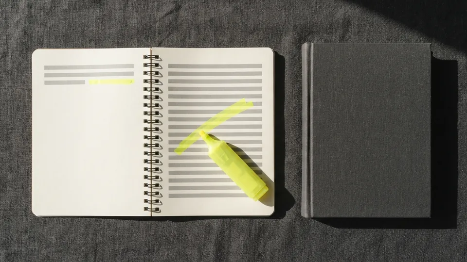 An open spiral notebook with a yellow line and a highlighter, next to a closed cloth-bound book