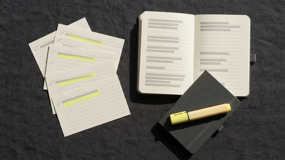 Index cards fanned beside an open notebook, each card with one yellow highlighter stroke and a pencilled tick in the margin