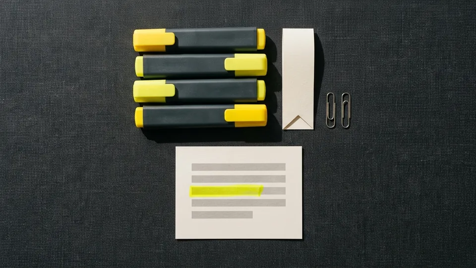Four highlighter pens lined up beside a paper bookmark, two paper clips and a card with one yellow stroke
